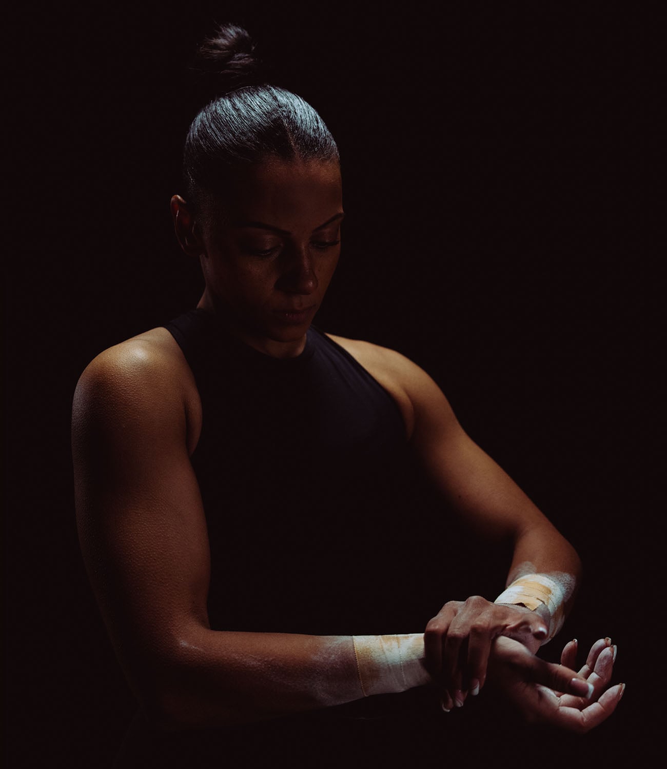 A woman in a black sleeveless top with her hair in a bun, shown in dramatic low light against a black background while applying white powder to her tapped wrists.
